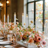 A centerpiece arrangement featuring tall white taper candles and floral accents on a wooden table.