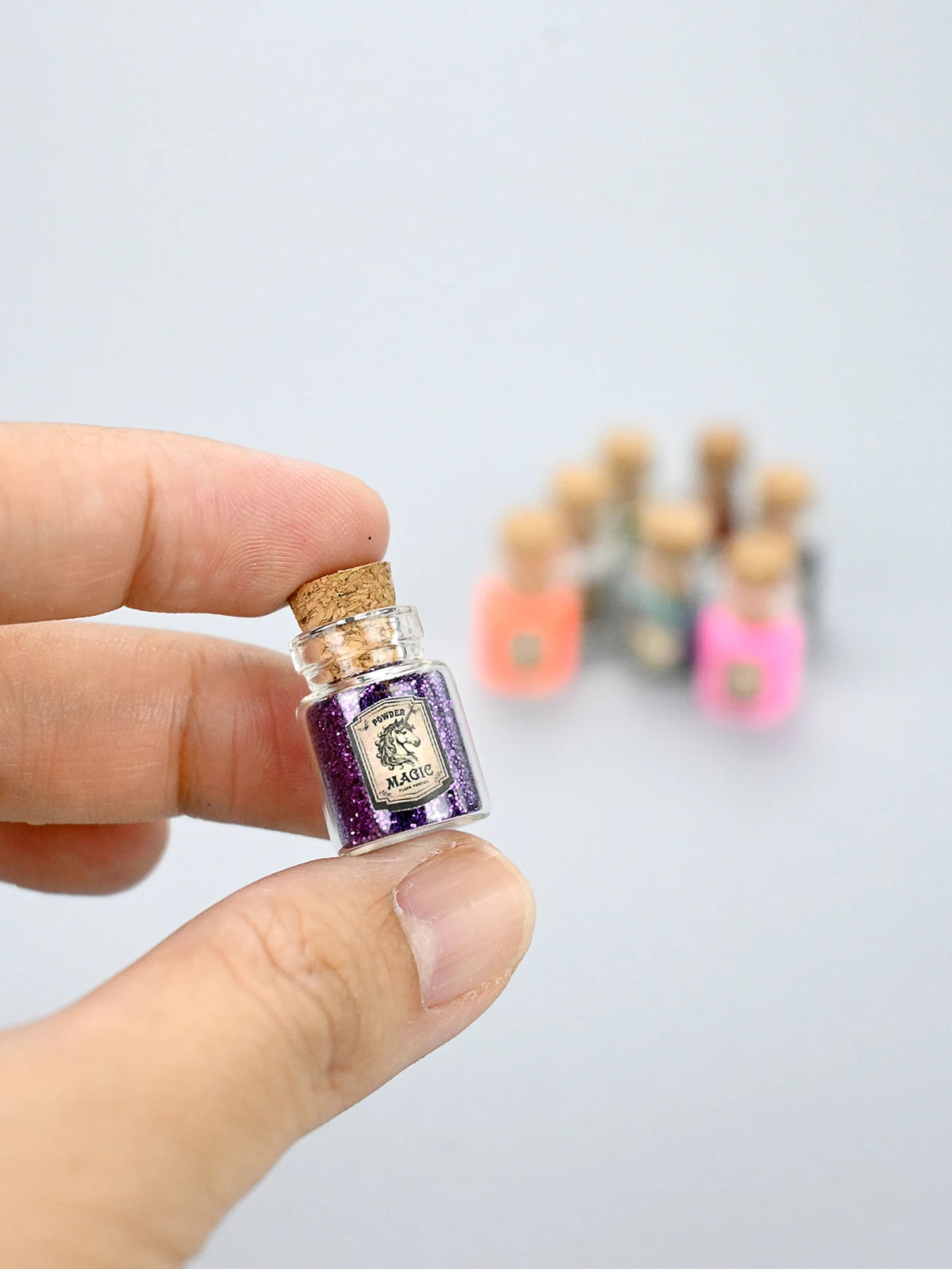 A hand holds a small glass bottle of purple glitter powder for party decorations next to several other similar bottles.