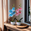 A pair of realistic artificial orchid potted plants, one blue and one pink, displayed on a wooden surface.