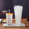 A glass of white beverage next to a clear plastic container filled with disposable drinking straws on a wooden table.