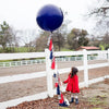 A girl stands in front of a white fence with a navy blue balloon floating above.