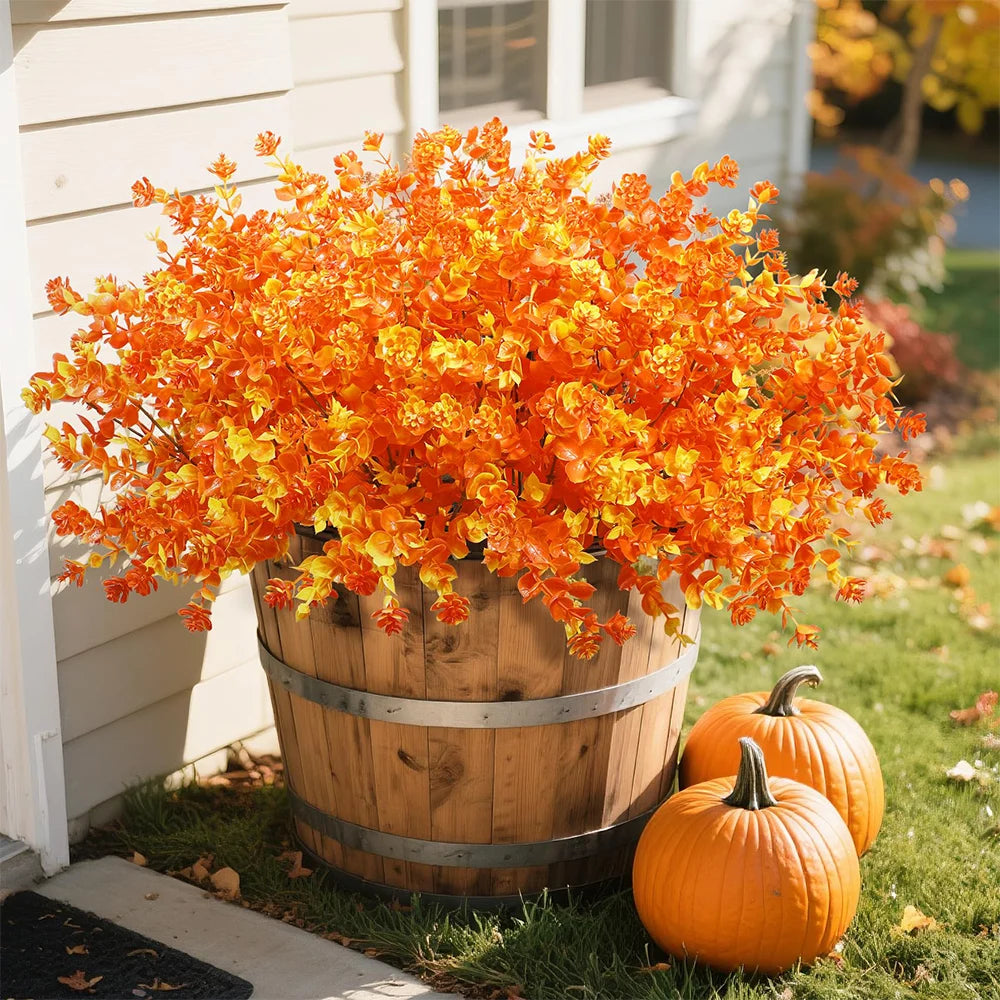 A vibrant display of orange and yellow artificial fall foliage in a wooden barrel planter with an orange pumpkin nearby.