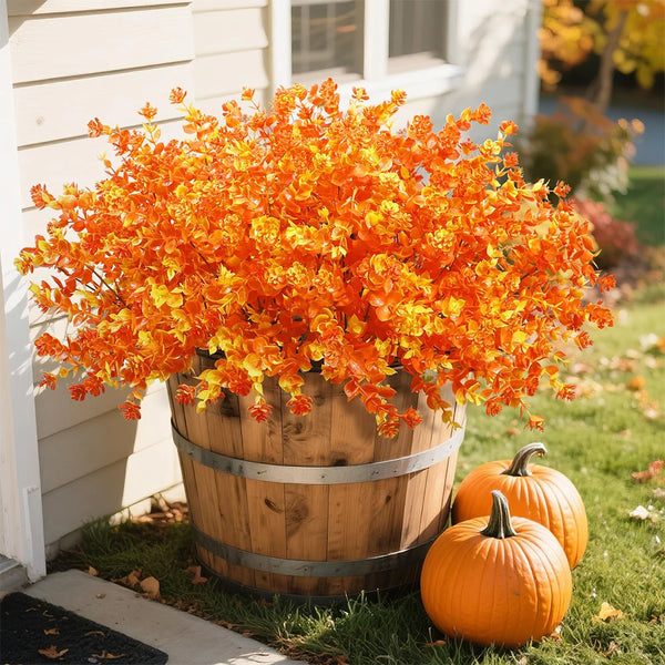 A vibrant display of orange and yellow artificial fall foliage in a wooden barrel planter with an orange pumpkin nearby.