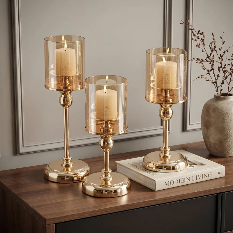 Three glass pillar candle holders with gold metal bases and candles on a dark wooden surface.