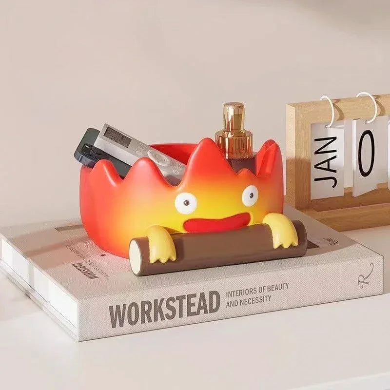 A resin cartoon fire person tray with an orange and yellow face sits on a book next to a wooden frame.