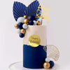 A round cake decorated with blue and white balls, gold accents, and a gold birthday topper on a white stand.