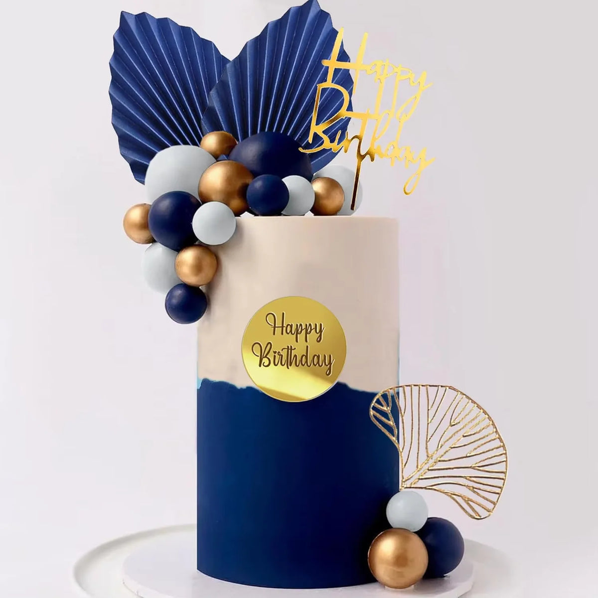 A round cake decorated with blue and white balls, gold accents, and a gold birthday topper on a white stand.