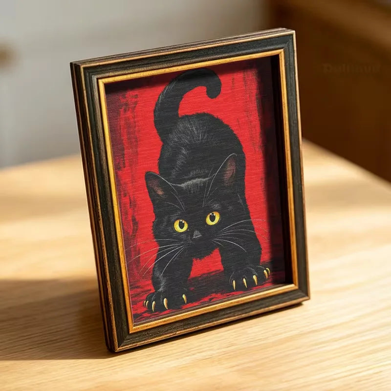A framed photograph of a black cat with yellow eyes against a red background, displayed on a wooden surface.