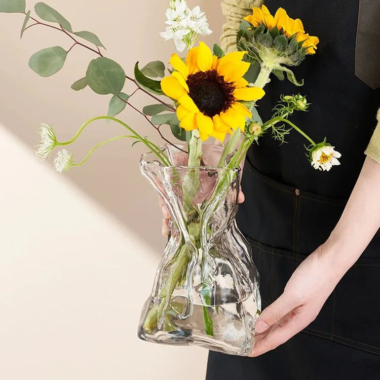A hand holds a clear glass vase shaped like a money bag filled with yellow and white flowers and green foliage.