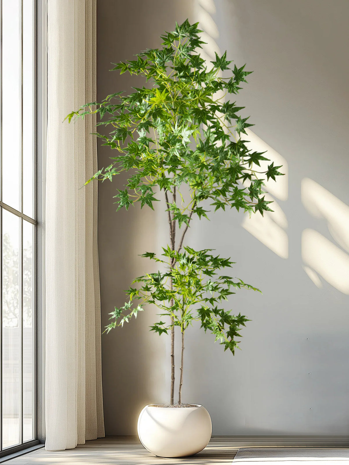 A tall artificial maple tree with silk green leaves is displayed in a white pot next to a window.