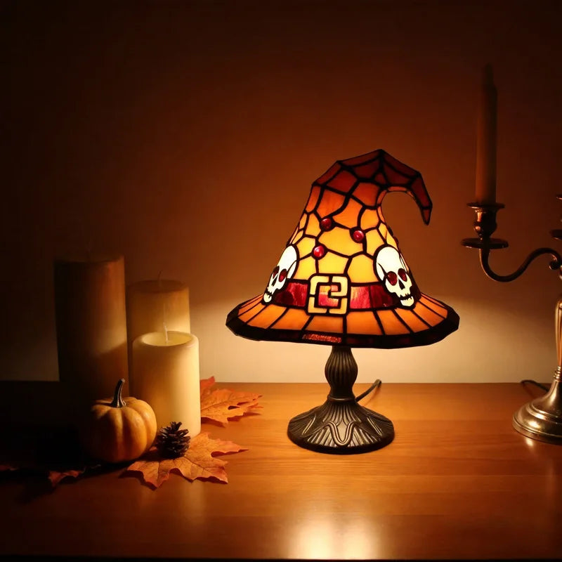 A witch hat night light with a stained glass effect illuminates a dark wooden surface next to candles and autumn leaves.
