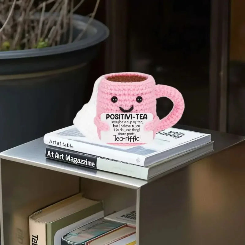 A pink handmade crochet tea cup ornament with a smiling face sits on a stack of books.
