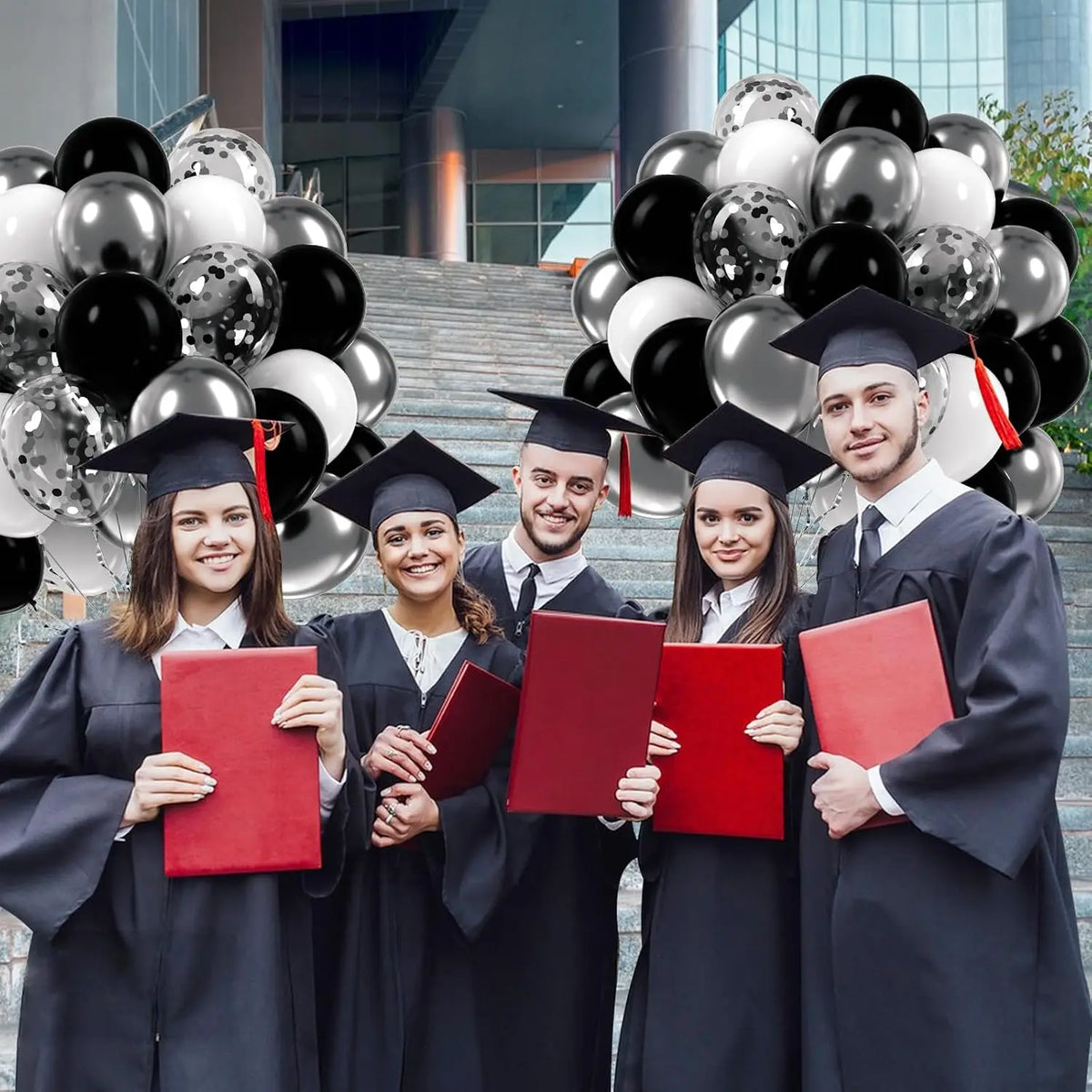 Five graduates in black gowns and caps hold red folders in front of a backdrop of silver and black balloons.