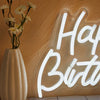 A white LED neon sign spelling out Happy Birthday is mounted on a textured wall next to a vase of yellow flowers.