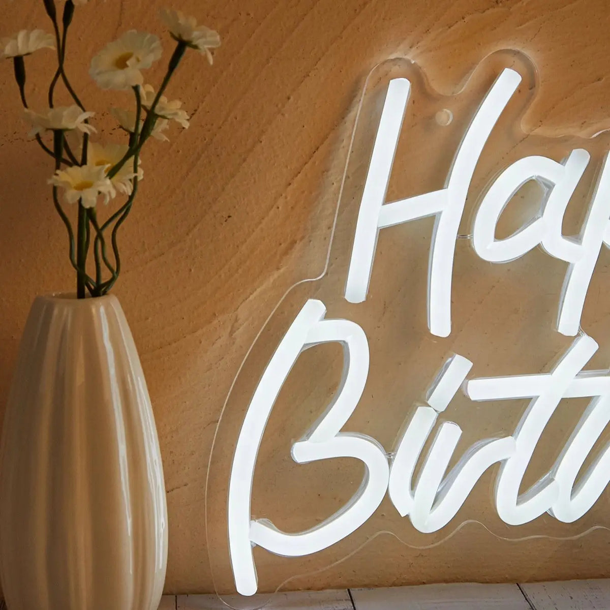 A white LED neon sign spelling out Happy Birthday is mounted on a textured wall next to a vase of yellow flowers.