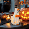 A white ghost figurine with an orange hat and carved features sits next to a glowing candle and a carved pumpkin.