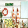A vintage metal wall sign reading WITCH WAY is mounted on a white wooden wall next to a red life preserver and striped towels