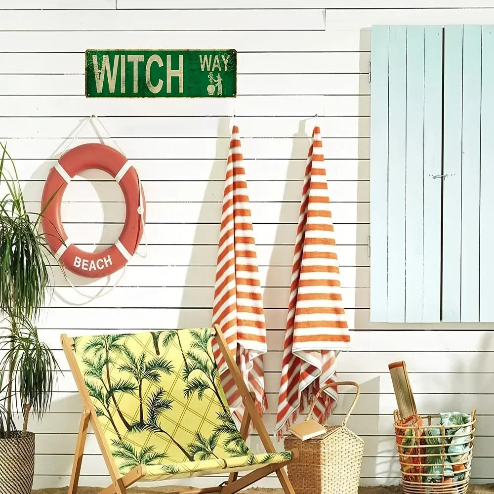 A vintage metal wall sign reading WITCH WAY is mounted on a white wooden wall next to a red life preserver and striped towels