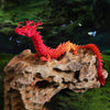 A vibrant red dragon figurine is perched on a rock amidst green moss, with small blue accents flying around it.