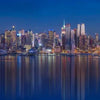 A panoramic night view of the New York City skyline reflected on the water under a deep blue sky.