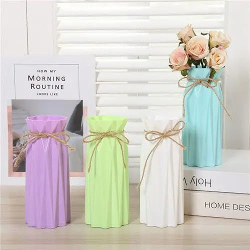 A modern origami plastic vase in light blue sits on a wooden surface next to a white striped blind.