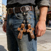 A person wearing denim jeans is holding a handcrafted walnut cross keychain with leather cord.