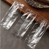 Three clear plastic disposable spoons packaged in individual sealed plastic sleeves on a wooden surface.