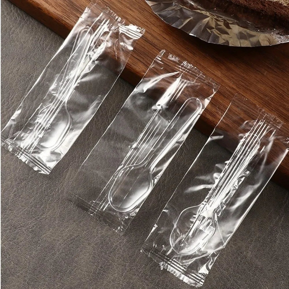 Three clear plastic disposable spoons packaged in individual sealed plastic sleeves on a wooden surface.