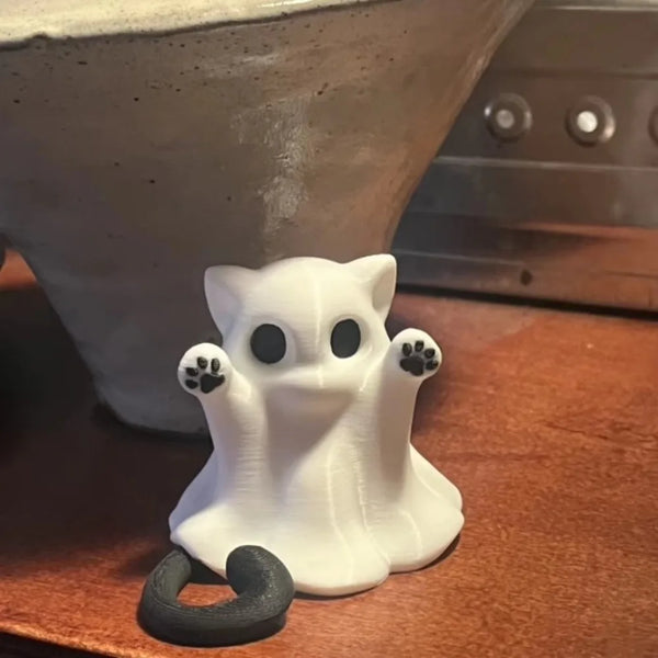 Una figura blanca de un gato fantasma con ojos y patas negras, sobre una superficie de madera junto a un cuenco de cerámica.