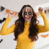 A woman wearing a full face horror costume mask resembling a creepy clown with exaggerated features and dark makeup.