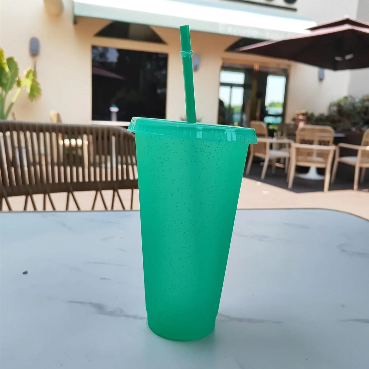 A bright green glitter party cup with a straw is placed on a white table outdoors.