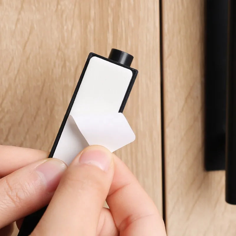 A hand is holding a white self-adhesive cabinet handle, demonstrating the no-drill pull mechanism.