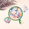 A portable foldable cooling handheld fan with a floral pattern is shown next to some dried purple flowers and a piece of pape