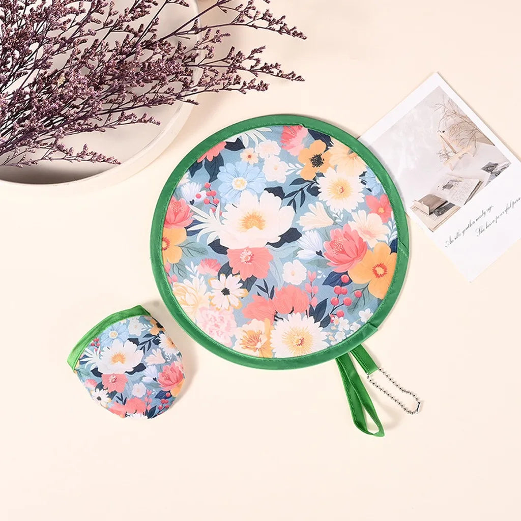 A portable foldable cooling handheld fan with a floral pattern is shown next to some dried purple flowers and a piece of pape