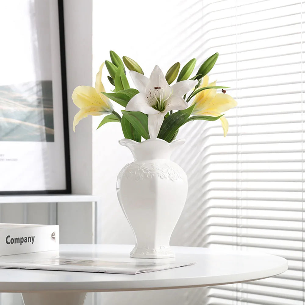 Elegant white ceramic vase holding yellow and white lilies on a white table near a window.