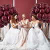 Three brides in white and blush gowns pose in front of a backdrop of burgundy red latex balloons.