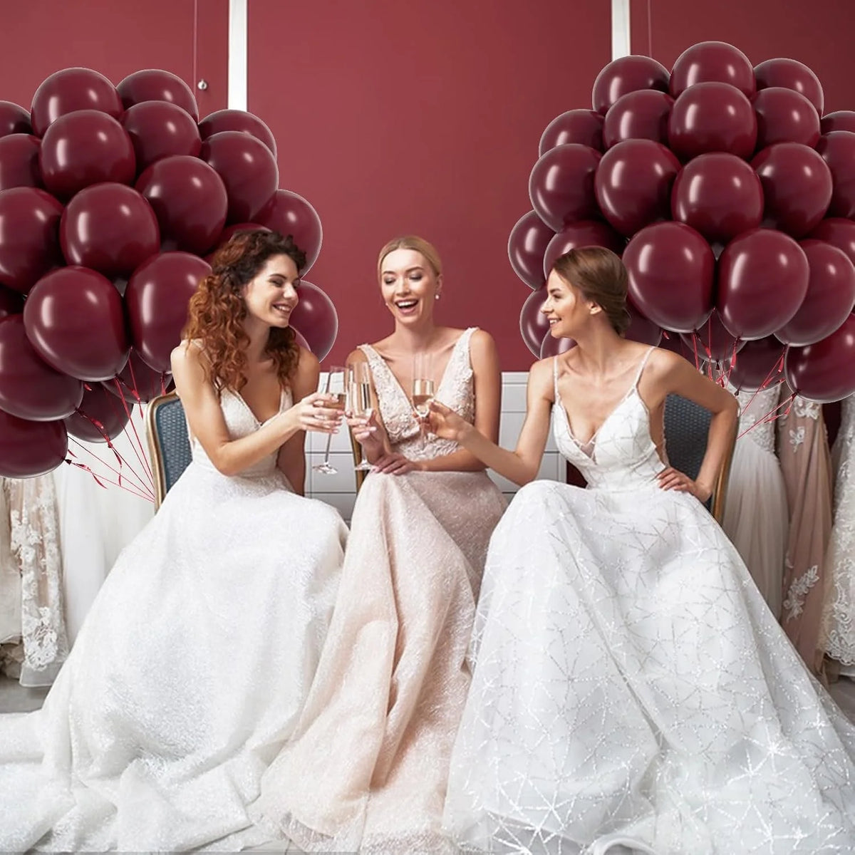 Three brides in white and blush gowns pose in front of a backdrop of burgundy red latex balloons.
