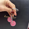 A hand holds a silver hoop and a pink cherry charm bag pendant with a keychain against a dark background.
