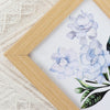 A watercolor floral painting of light blue flowers framed in natural wood.