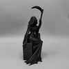 A black figurine of a gothic female grim reaper holding a large scythe.