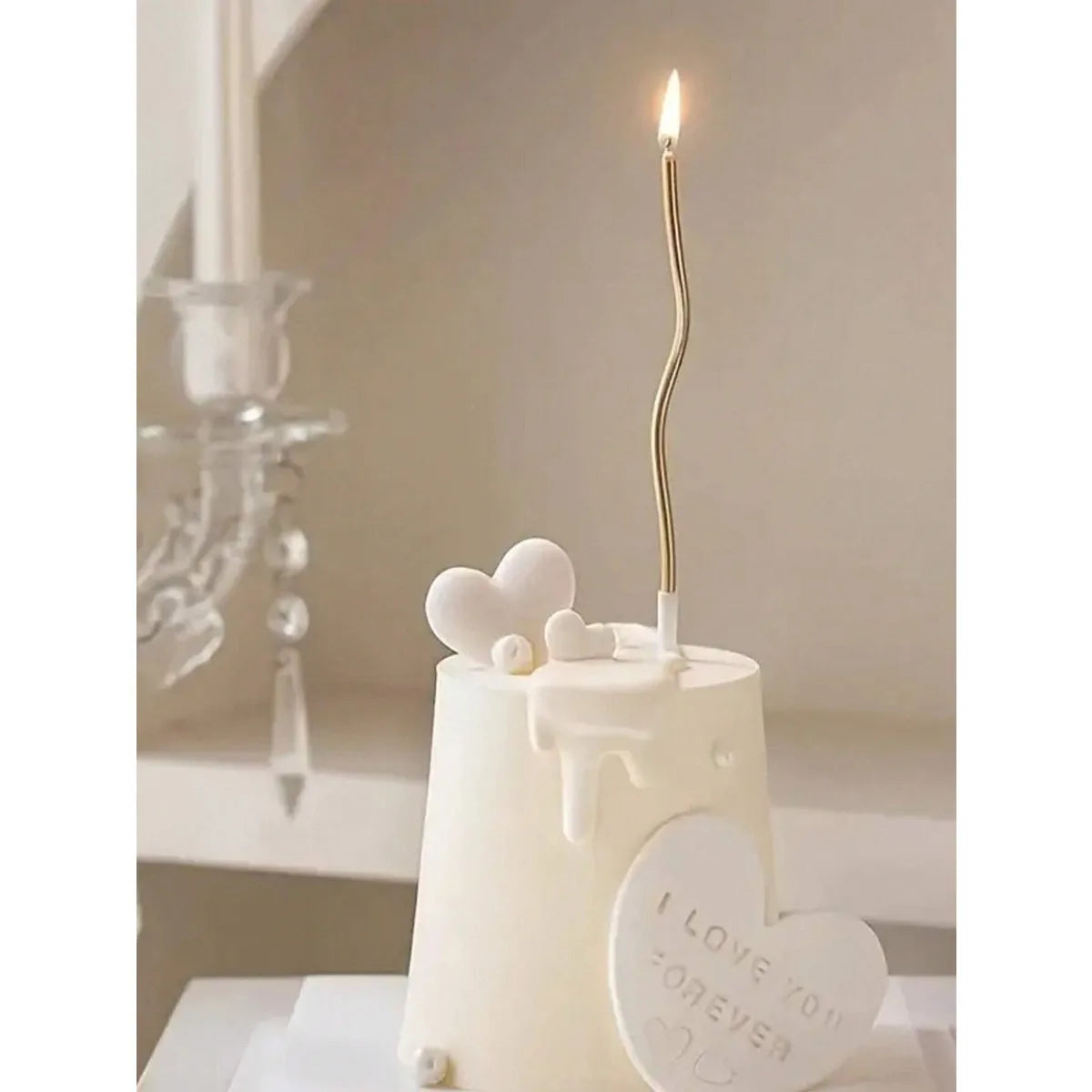 A white cake topped with heart decorations and a tall golden candle burning on a light surface.