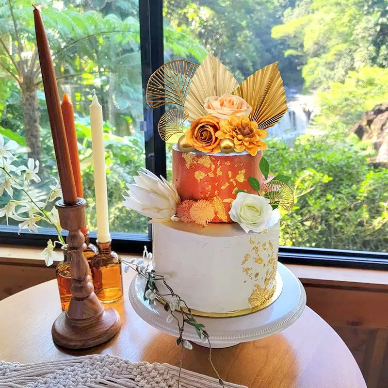 A white cake decorated with gold leaf accents and orange flowers, set against a blurred green outdoor background.