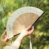 A person holds an elegant bamboo folding fan with a tassel against a blurred green outdoor background.