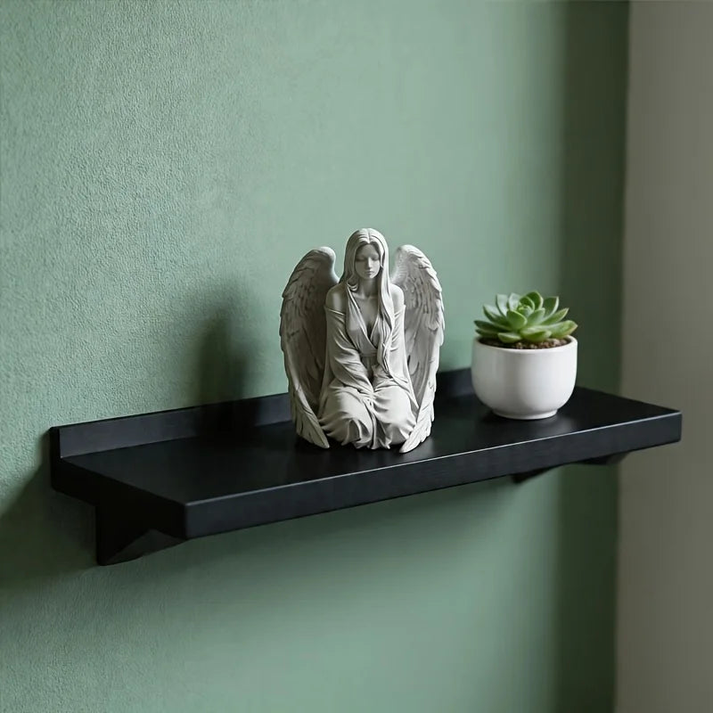 Estatua de ángel de mármol blanco con alas sobre un estante negro contra una pared verde apagada.