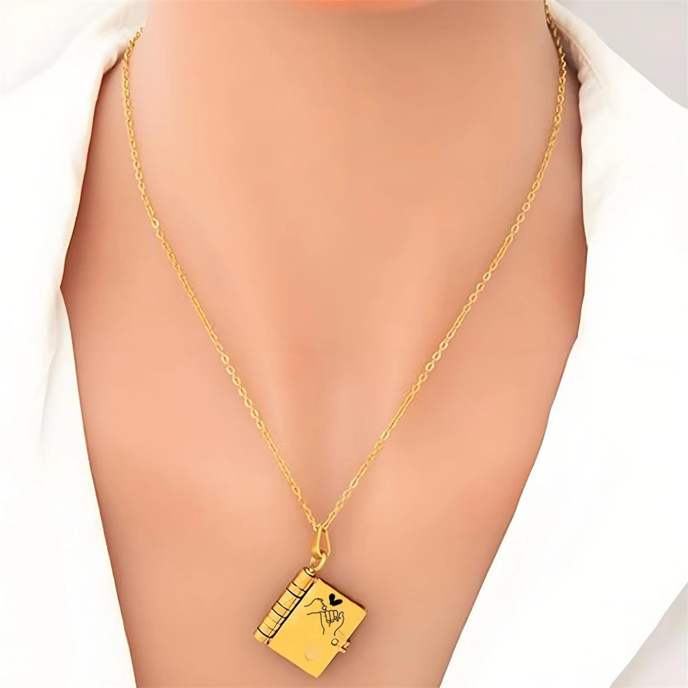 A gold chain necklace with an openable book pendant engraved with a message, worn against light skin.
