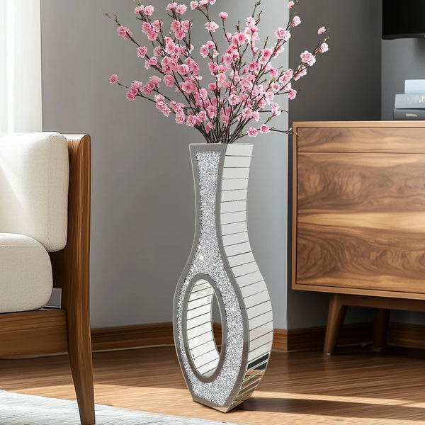 A tall silver mirrored vase with a crushed diamond finish holds pink cherry blossoms on a wooden cabinet.