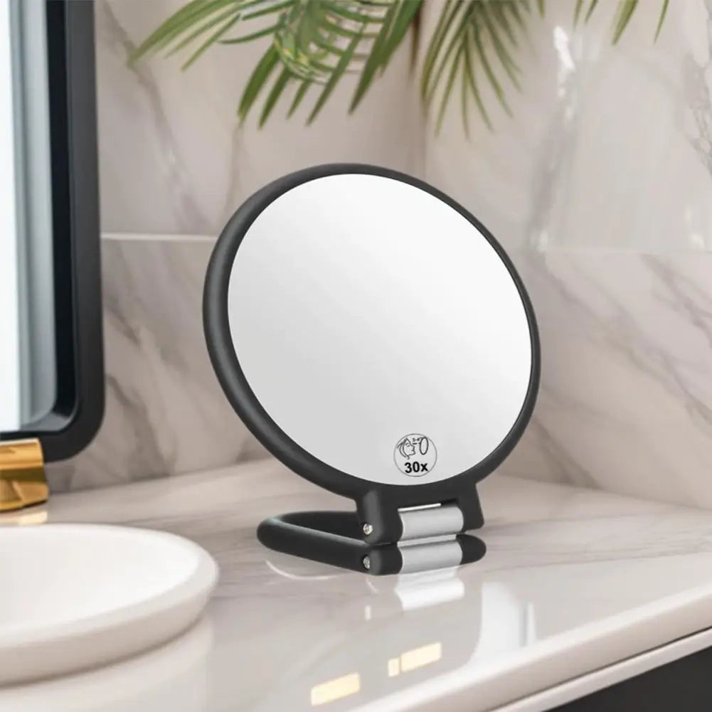 A round, black handheld mirror with a silver handle and 30x magnification is placed on a white countertop.