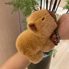 A fluffy brown capybara animal wristband and ring is displayed on a person's hand against a blurred green plant background.