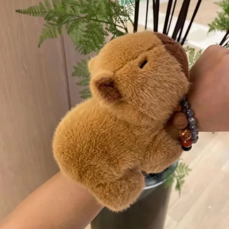 A fluffy brown capybara animal wristband and ring is displayed on a person's hand against a blurred green plant background.