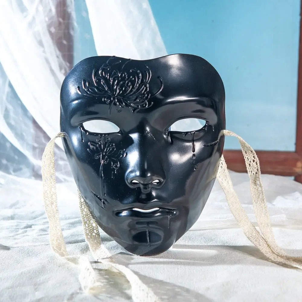 A black ancient style masquerade mask with intricate designs and light colored ties resting on white fabric.